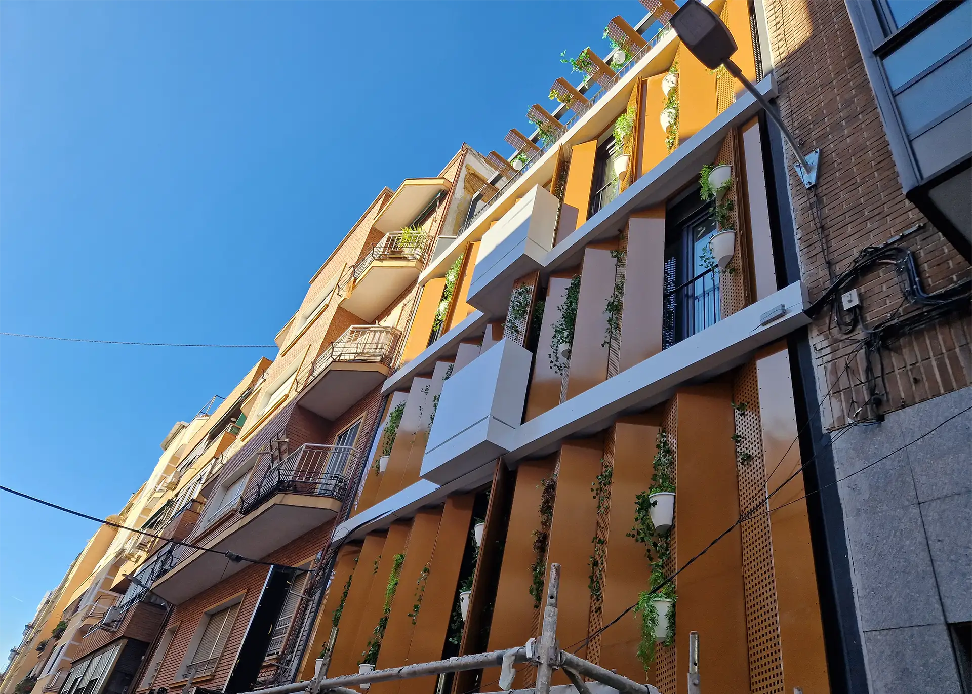 Perspectiva desde la base de la fachada del edificio en Tetuán, Madrid. Los paneles perforados, combinados con macetas de plantas, destacan el diseño contemporáneo y sostenible.