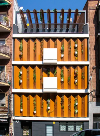 Jeronima10-Exterior1 Fachada de un moderno edificio residencial con paneles verticales naranjas y plantas colgantes en las paredes.