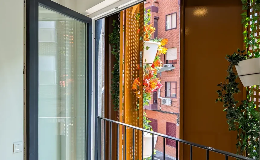 Vista desde el interior de un apartamento a través de la puerta abierta de un balcón, mostrando las lamas exteriores y la calle.